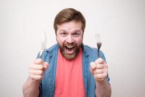 hongerig Kaukasisch Mens met een vork en een mes in handen, looks met gek ogen Bij de camera en lacht uit luid. eetpatroon concept. foto