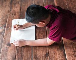 jongen zittend Aan de houten verdieping schrijven een boek of aan het doen huiswerk Bij huis. top visie van kind schrijven boek foto