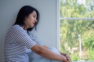 een Aziatisch vrouw za alleen in de huis met haar knieën naast de venster. met verdrietig en teleurgesteld gezichten en psychologisch symptomen. depressie of een niet verwacht zwangerschap concept. foto