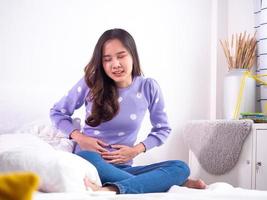Aziatisch Dames met menstruatie en menstruatie- krampen. een meisje zittend in pijn in een bed Bij huis. foto