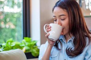een Aziatisch vrouw in een mooi zo humeur is drinken koffie gelukkig. Dames voelen ontspannen met de aroma van een drinken in de huis of cafe. voedsel en drinken Aan een helder ochtend- foto