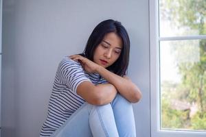 een Aziatisch vrouw zitten alleen in de huis met haar knieën naast de venster. met verdrietig en teleurgesteld gezichten en psychologisch symptomen. depressie of een niet verwacht zwangerschap concept. foto
