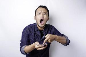 geschokt Aziatisch Mens vervelend blauw overhemd en Holding zijn telefoon, geïsoleerd door wit achtergrond foto