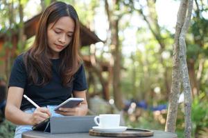 Aziatisch vrouw op zoek Bij verdiensten Aan haar telefoon Bij een koffie winkel waar ze kan Gaan naar werk. temidden van groen natuur foto
