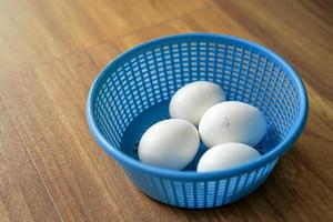 4 witte kippeneieren in blauwe plastic mand op houten vloer. 1 van de 4 kip is klaar om de eierschaal van binnenuit te breken. foto