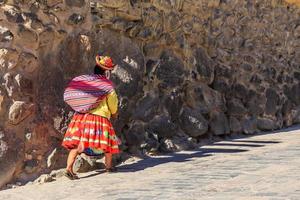 Peruaanse vrouw cholita gekleed in traditioneel kleurrijk lap, draag- de zak en wandelen omhoog de straat met steenachtig muren, inkan heilig vallei, ollantaytambo, Peru foto