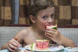 een vrolijk jongen eet watermeloen Bij huis Bij de tafel, huis comfort, de warmte van de familie haard foto