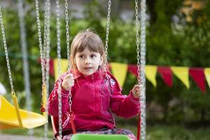 een weinig meisje in een rood jasje zit Aan een keten carrousel rijden. foto