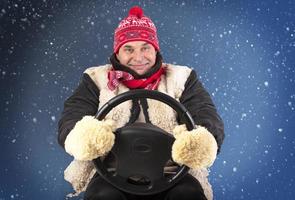 grappig volwassen Mens in een hoed en vacht wanten houdt een auto stuurinrichting wiel Aan een blauw besneeuwd achtergrond. foto