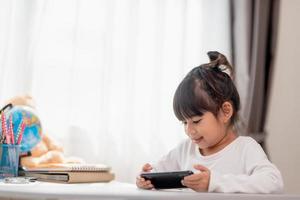 Aziatisch weinig meisje gebruik makend van een telefoon dichtbij omhoog, afgeleid van aan het studeren, zittend Bij een tafel met notitieboekjes, een mooi kind hebben pret met een smartphone, aan het kijken de webinar, thuisonderwijs foto