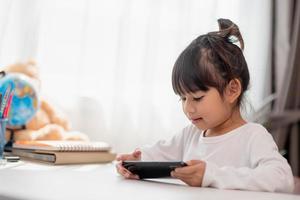 Aziatisch weinig meisje gebruik makend van een telefoon dichtbij omhoog, afgeleid van aan het studeren, zittend Bij een tafel met notitieboekjes, een mooi kind hebben pret met een smartphone, aan het kijken de webinar, thuisonderwijs foto