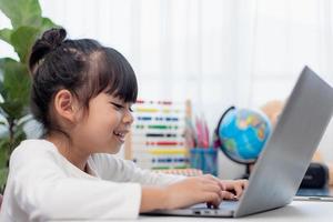Aziatisch schoolmeisje aan het doen haar huiswerk met laptop Bij huis. kinderen gebruik gadgets naar studie. onderwijs en afstand aan het leren voor kinderen. thuisonderwijs gedurende quarantaine. blijven Bij huis foto