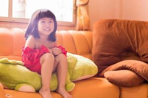 Aziatisch kinderen zijn schattig of kind meisje en kleuterschool leerling hebben gelukkig glimlacht wit tanden en lach met vervelend een rood jurk voor mode in de leven kamer. foto