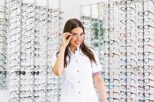 aantrekkelijk jong vrouw dokter in oogheelkunde kliniek. dokter oogarts is staand in de buurt schappen met verschillend bril. foto