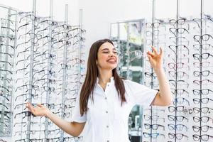 poseren optometrist vrouw in bril op te slaan glimlachen op zoek Bij camera. portret van jong glimlachen vrouw staand in voorkant van kleinhandel Scherm in optisch winkel foto