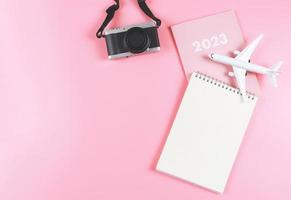 vlak leggen van blanco papier Notitie boek Aan roze dagboek 2023 met vliegtuig model- en camera Aan roze achtergrond. reizen plan concept. foto