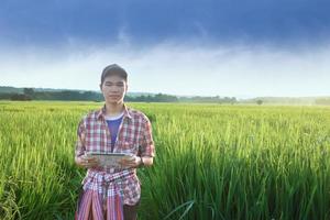 jong Aziatisch tiener jongen in plaid shirt, draagt pet en Holding tablet in handen, staand en gebruik makend van zijn tablet naar enquête informatie van rijst- groeit en naar Doen school- project werk in rijst- rijstveld veld. foto