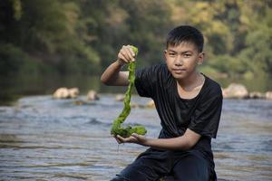 Aziatisch schooljongen Holding zoetwater algen van duiken in de rivier- en trekken het omhoog naar studie de vruchtbaarheid van de rivieren natuur en inclusief naar Doen zoetwater algen in zijn milieu project werk. foto