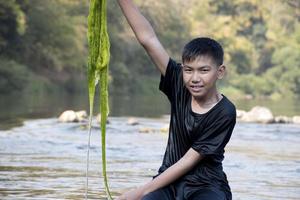 Aziatisch schooljongen Holding zoetwater algen van duiken in de rivier- en trekken het omhoog naar studie de vruchtbaarheid van de rivieren natuur en inclusief naar Doen zoetwater algen in zijn milieu project werk. foto