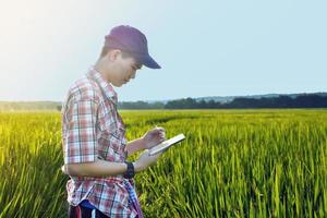 jong Aziatisch tiener jongen in plaid shirt, draagt pet en Holding tablet in handen, staand en gebruik makend van zijn tablet naar enquête informatie van rijst- groeit en naar Doen school- project werk in rijst- rijstveld veld. foto