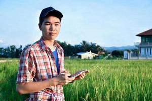 jong Aziatisch tiener jongen in plaid shirt, draagt pet en Holding tablet in handen, staand en gebruik makend van zijn tablet naar enquête informatie van rijst- groeit en naar Doen school- project werk in rijst- rijstveld veld. foto