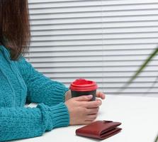 een vrouw in een blauw trui houdt een papier kop met koffie Bij een wit tafel. koffie breken foto