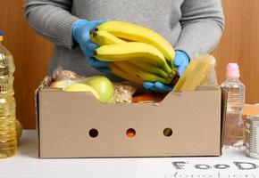 vrouw in handschoenen houdt verzamelen voedsel, fruit en dingen en een karton doos voor helpen die in nodig hebben foto