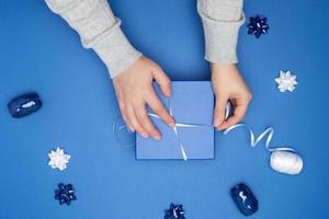 twee vrouw handen in een grijs trui houden Gesloten plein blauw doos met een boog Aan een blauw klassiek achtergrond foto