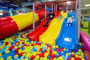 gelukkig kinderen spelen Bij binnen- Speel centrum speelplaats. kinderen dia's in gekleurde glijbaan in ballen in bal zwembad. foto