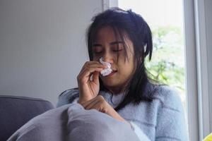 Aziatisch vrouw is ziek hebben benauwd neus, vloeiende water in de handen, afvegen papier, hoog koorts zittend Aan de sofa binnen de huis foto