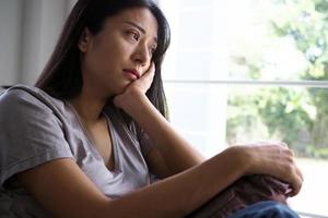 Aziatisch vrouw zittend binnen de huis op zoek uit Bij de venster. vrouw verward, teleurgesteld, verdrietig en van streek foto