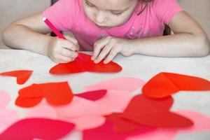 kind trekt met een rood viltstift pen Aan eigengemaakt papier gesneden harten, voorbereidingen treffen voor de vakantie moeder dag, Valentijnsdag dag. diy vakantie kaart met een rood papier hart, een symbool van liefde. foto