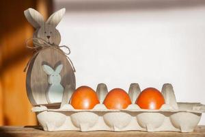 Aan een houten achtergrond in een bord zijn drie Pasen rood eieren geschilderd met natuurlijk kleuren en een Pasen konijn in de stralen van de helder zon, minimalisme, kopiëren ruimte. voorbereidingen treffen naar vieren Pasen foto