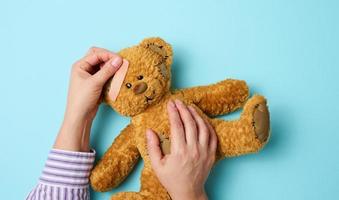 vrouw hand- houdt een bruin teddy beer en lijmen een medisch Zelfklevend gips Aan een blauw achtergrond, tram behandeling foto