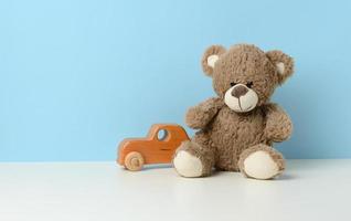 schattig bruin teddy beer zit Aan een wit tafel en een houten kinderen speelgoed- auto, blauw achtergrond foto