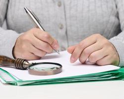 vrouw zit Bij een wit tafel en houdt een metaal pen over- een stapel van papieren, in de andere hand- een vergroten glas foto