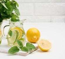 transparant glas karaf met plakjes van citroen, limoen en munt bladeren Aan een wit tafel, ontgiften. achter de ingrediënten voor de drinken foto