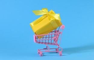 geel geschenk doos met een zijde boog in een miniatuur metaal trolley Aan een licht blauw achtergrond foto