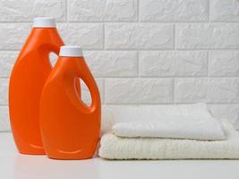 twee oranje plastic flessen van vloeistof wasmiddel en een stack van handdoeken Aan een wit plank, huis wassen foto