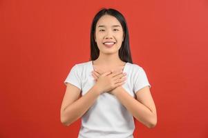 portret van gelukkig jong Aziatisch vrouw in blauw overhemd tonen haar hart gevulde met liefde en Holding handen Aan haar borst geïsoleerd Aan rood achtergrond foto