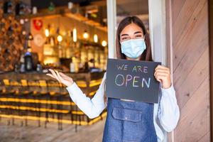 gelukkig vrouw serveerster met beschermend gezicht masker Holding Open teken terwijl staand Bij cafe of restaurant deuropening, Open opnieuw na slot naar beneden ten gevolge naar het uitbreken van coronavirus covid-19 foto