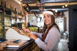 serveerster vervelend een schort staand door een punt van uitverkoop terminal en lachend terwijl werken in een restaurant. mooi vrouw vervelend gezicht schild gedurende coronavirus pandemisch staand door contant geld registreren foto