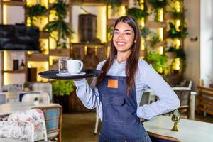 jong vrouw werken Holding een dienblad met koffie en glas van water. mooi serveerster vervelend schort. portret van een glimlachen serveerster Holding dienblad van drankjes in restaurant foto