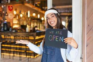 serveerster met gezicht schild groet klanten in een restaurant of een caffe met fles van alcohol naar desinfecteren hun handen voordat binnenkomen. jong vrouw Holding teken wij zijn Open foto