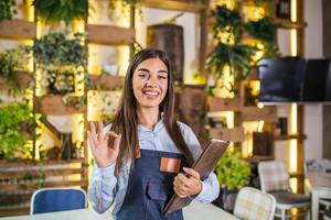 mooi vrouw serveerster glimlachen in de restaurant Holding een menu en tonen OK teken. foto