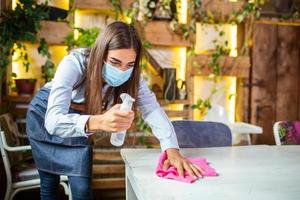 portret van aantrekkelijk vrouw serveerster vervelend gezicht masker en schoonmaak de tafel met alcohol en nat doekjes voordat Welkom klant. nieuw normaal met coronavirus het uitbreken hygiëne restaurant concept. foto