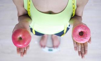 vrouw meten lichaam gewicht Aan weging schaal Holding donut en appel. snoepgoed zijn ongezond rommel voedsel. diëten, gezond aan het eten, gewicht verlies. zwaarlijvigheid. foto