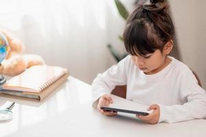 Aziatisch weinig meisje gebruik makend van een telefoon dichtbij omhoog, afgeleid van aan het studeren, zittend Bij een tafel met notitieboekjes, een mooi kind hebben pret met een smartphone, aan het kijken de webinar, thuisonderwijs foto