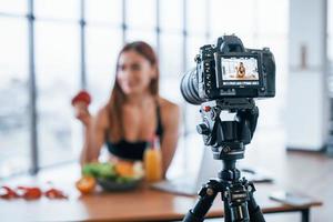 vrouw vlogger met sportief lichaam staand binnenshuis in de buurt tafel met gezond voedsel foto