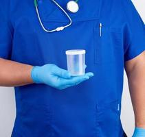 dokter in blauw uniform en latex handschoenen is Holding een leeg plastic houder voor nemen urine monsters foto